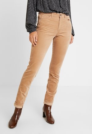 Kobieta ubrana w beżowe sztruksowe spodnie ze podwiniętymi nogawkami, czarną wzorzystą bluzkę z długim rękawem oraz brązowe botki na obcasie, stojąca na białej podłodze.