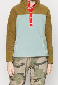 Jasnozielony polar z brązowym kołnierzem, czerwonym zapięciem na guziki i dwiema bocznymi kieszeniami. Materiał jest miękki z teksturowanym wzorem.