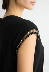 Top corto nero a maniche corte con perline decorative e paillettes lungo la cucitura della spalla, che offre un dettaglio strutturato e decorato.