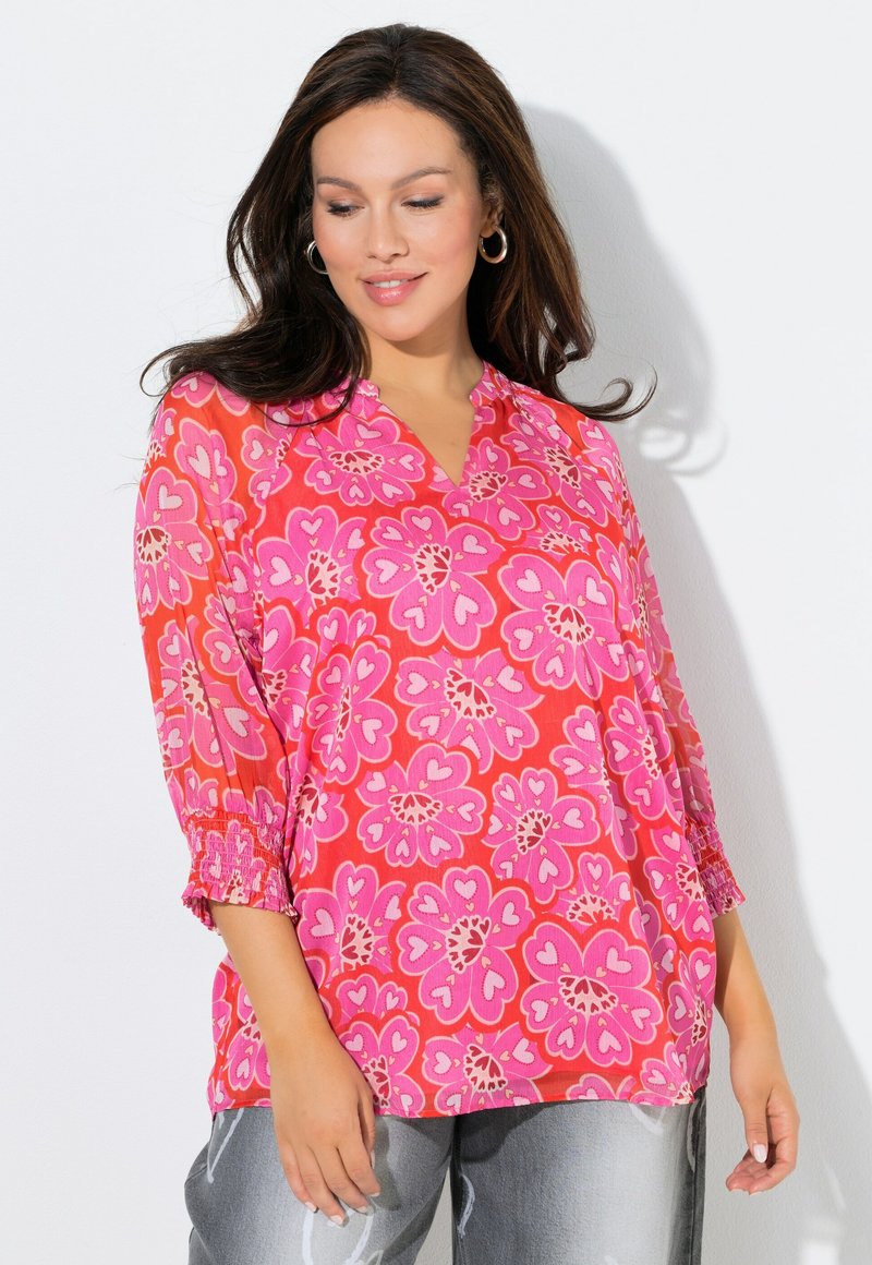 Różowa kwiatowa bluzka z dekoltem w kształcie litery V, rękawami do połowy przedramienia i marszczonymi mankietami. Ozdobiona dużymi, sercowatymi kwiatami w różnych odcieniach różu.