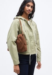 Μοντέρνο ανοιχτοπράσινο μπουφάν με κουκούλα και κορδόνια, με τσέπη στο μπροστινό μέρος, συνδυασμένο με καφέ σουέτ τσάντα. Φοράει σκούρο τζιν παντελόνι. Υφασμάτινη υφή και casual σχεδιασμός.