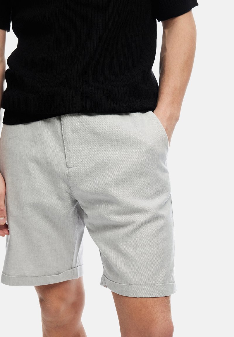 Man in een lichtgrijze short met opgerolde zoom, met een hand in zijn zak, en een zwart T-shirt met korte mouwen, tegen een witte achtergrond.