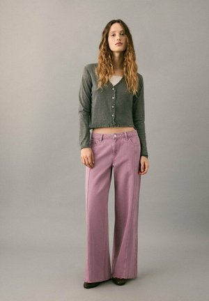 Graue, kurz geschnittene Strickjacke mit Knöpfen, kombiniert mit hoch taillierten, weit geschnittenen pinken Jeans. Modell steht vor einem neutralen Hintergrund.