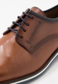 Chaussure en cuir marron avec une finition lisse, dotée de lacets noirs, de détails de couture, d'un bout arrondi et d'une semelle rayée blanche et marine.