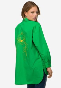 Camicia oversize verde realizzata in tessuto morbido, con una grafica ricamata gialla sul retro. Maniche lunghe con polsini abbottonati.