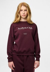 Młoda kobieta o długich blond włosach, ubrana w bordową bluzę i spodnie, na bluzie napis: "Aesthetic Club established 1892."