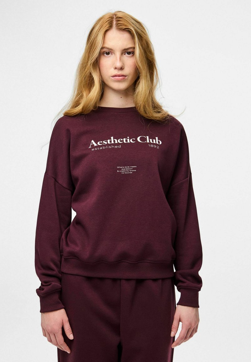 Młoda kobieta o długich blond włosach, ubrana w bordową bluzę i spodnie, na bluzie napis: "Aesthetic Club established 1892."