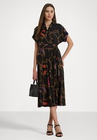 Čierne obopnuté šaty s krátkymi rukávmi, s vzorom zlatých a farebných reťazcov; doplnené malou čiernou kabelkou a sandálmi s remienkami.