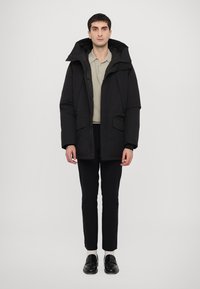 Parka neagră căptușită, cu guler înalt, închidere cu fermoar, buzunare frontale mari și glugă ajustabilă. Purtați peste un tricou polo de culoare deschisă.