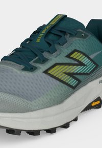 Trailrunning-Schuh in Türkis und Grau mit strukturierter Sohle, blauen Schnürsenkeln, gelb-grünem New Balance-Logo und dem Label „FuelCell rebel trail“ auf der Zunge.