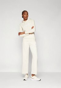 Krēmkrāsas kapuci džemperis ar baltu logotipu, kopā ar saskaņota krēmkrāsas platajiem sporta biksēm un baltiem sporta apaviem, ar gadījuma un relaksētu dizainu.