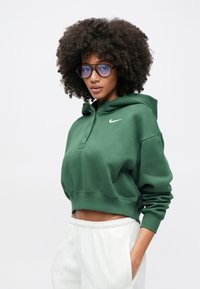 Sweat à capuche vert raccourci avec cordon de serrage à la capuche, orné d'un petit logo Nike blanc. Le modèle porte de grosses lunettes bleues et un pantalon de couleur claire.