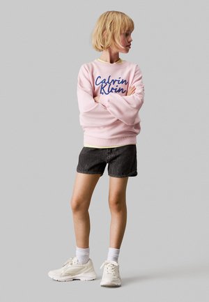 Różowy sweatshirt z niebieskim haftowanym logo "Calvin Klein", połączony z ciemnoniebieskimi szortami dżinsowymi i białymi trampkami. Model ma krótkie blond włosy.