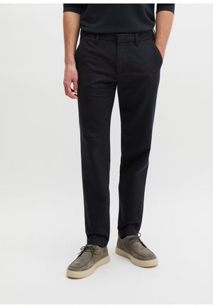 Man die zwarte slim-fit broek en grijze suède veterschoenen draagt, staand met één hand in zijn zak.