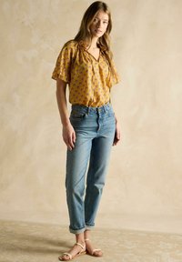 Kollane lillemustriga pluus lühikeste lokkis varrukatega, kombineerituna helesiniste kõrge vöökohaga teksade ja beežide sandaalide kingadega.