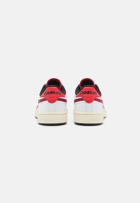 Hvide sneakers med sorte og røde accenter, der har en glat læderoverdel, polstret krave og struktureret gummisål. Branding på hælen.