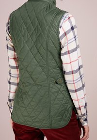 Pikowana zielona kamizelka z błyszczącej tkaniny, z wysokim kołnierzem, zapięciem na guziki z boku i dopasowanym krojem noszona na koszuli w kratę.