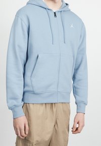 Ljusblå zip-up hoodie av mjukt tyg, med en känguruficka, dragsko i huvan och vit logotyp på vänster bröst.