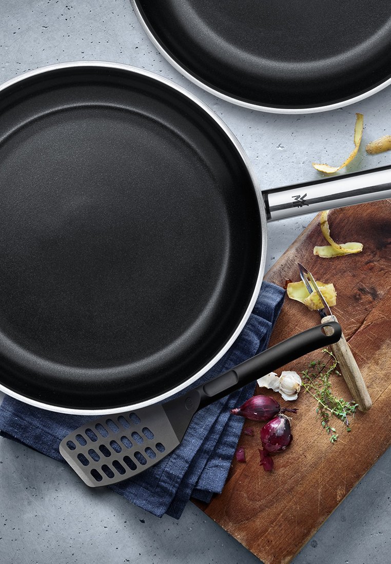 Nieprzywierająca czarna patelnia z srebrnym brzegiem, czarną rączką, w towarzystwie czarnej łopatki z otworami, ziołami, czosnkiem i czerwoną cebulą na drewnie.