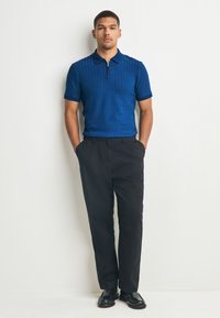 Blauw poloshirt met een gestructureerd geometrisch patroon, gecombineerd met een zwarte broek. Het shirt heeft een kraag en een halve ritssluiting bij de hals. Zwarte schoenen.