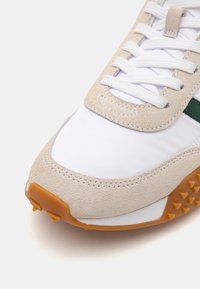 Beige en witte sneaker met suède en textielmaterialen, een textuurzool met rubberen noppen en contrasterende groene accenten op de bovenkant.