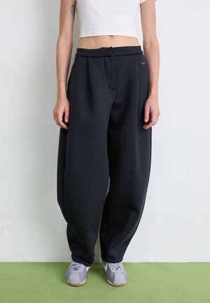 Svarte, løstsittende joggebukser laget av mykt stoff, med frontfold og sidelommer. Båret med grå sneakers på et grønt underlag.
