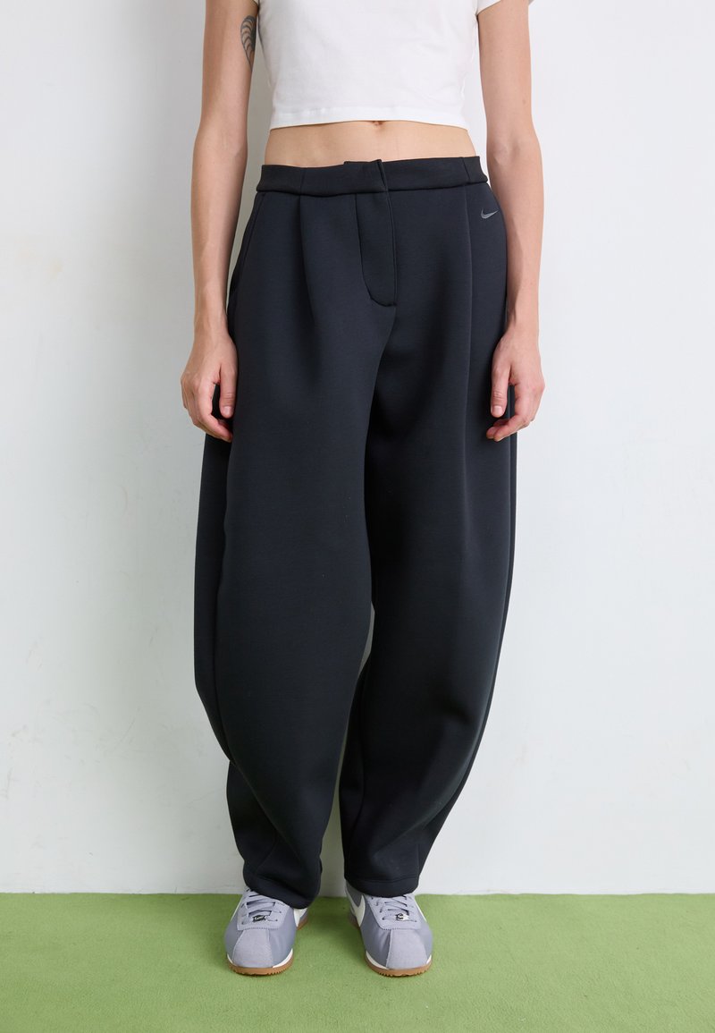 Pantalon de jogging noir ample en tissu doux, avec un pli avant et des poches latérales. Porté avec des baskets grises sur une surface verte.