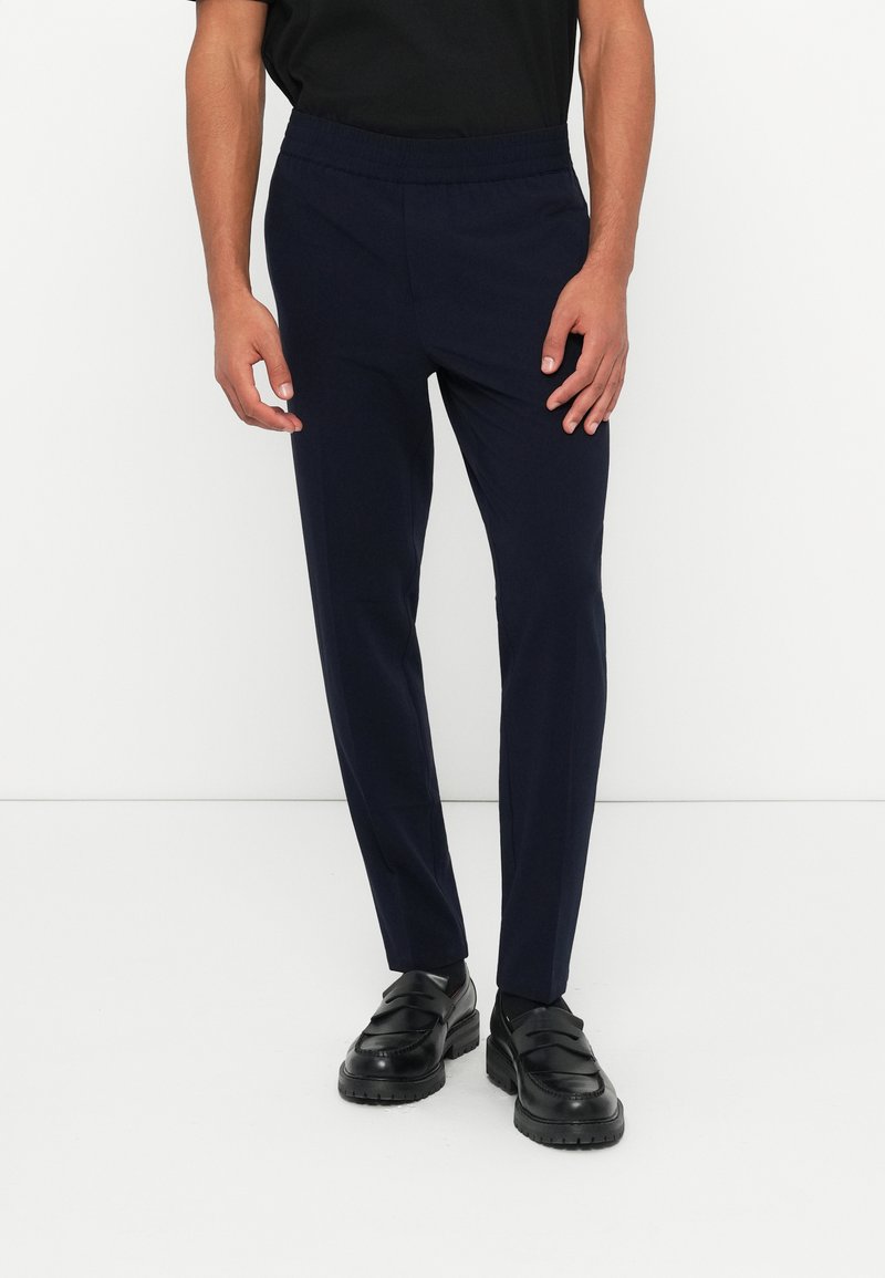 Calças azul-marinho ajustadas com um cós elástico, feitas de um tecido suave. Usadas com sapatos pretos de slip-on, apresentando um design minimalista.