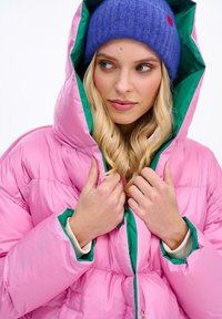 Růžová péřová bunda se zeleně podšitou kapucí, lesklým povrchem a prošitým vzorem. Modrá pletená čepice doplňuje vzhled.