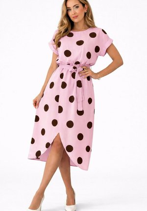 Femme en robe midi rose à grands pois marron foncé, manches courtes, ceinture à la taille, fente sur le devant et chaussures blanches à talons hauts.