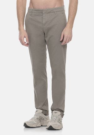 Uomo con pantaloni beige slim-fit e sneakers New Balance grigio chiaro, in piedi davanti a uno sfondo bianco uniforme.