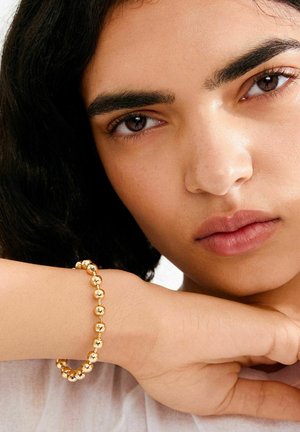 Zlatna narukvica s perlicama glatke površine, sadrži okrugle metalne perle različitih veličina, nošena na zapešću na neutralnoj pozadini.