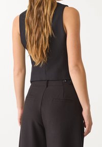 Czarny bezrękawnik w połączeniu z czarnymi spodniami z wysokim stanem. Gładka tkanina, dopasowany krój, guziki na tylnej stronie pasa.