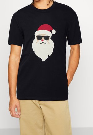 Person trägt ein schwarzes T-Shirt mit einem Grafikdruck von Santa Claus Gesicht, der eine Sonnenbrille und einen roten Hut trägt, kombiniert mit beigen Shorts.