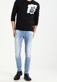 Svart sweatshirt med grafisk design, kombinerad med ljusblå slitna jeans. Modell som bär svarta ankelstövlar. Avslappnad outfit-presentation.