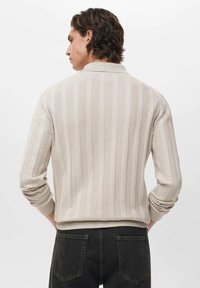 Suéter de punto a rayas beige con cuello, puños y dobladillo acanalados, que presenta una tela texturizada y un corte holgado, usado sobre pantalones oscuros.