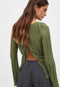 Grøn plisseret bluse med en åbning bagpå og bindebånd, lange ærmer og en afslappet pasform, parret med kulgrå bukser.