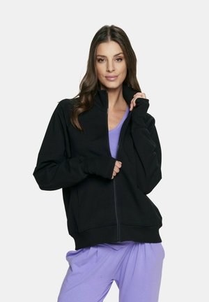 Czarna kurtka z zamkiem na wysokim kołnierzu, z prążkowanymi mankietami i dwoma przednimi kieszeniami; noszona na fioletowej bluzce o miękkiej, gładkiej teksturze.