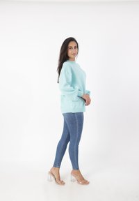 Maglione oversized verde menta con lunghe maniche a palloncino, abbinato a jeans blu aderenti e sandali con tacco trasparente, su uno sfondo bianco semplice.