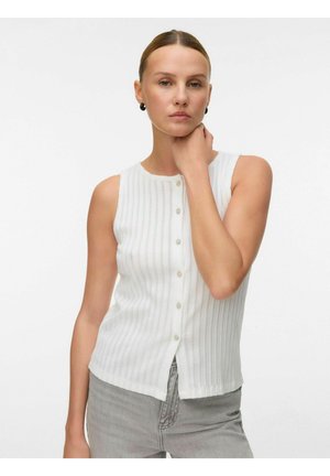 Vrouw met naar achteren gekamd haar, draagt zwarte oorbellen, een mouwloos wit topje met knopen en lichtgrijze broek, poseert met hand op de nek.