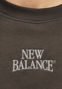 Εγγύς λήψη σκούρας γκρι φούτερ με στρογγυλή λαιμόκοψη και λευκή κεντητή επιγραφή "NEW BALANCE" στο κέντρο του στήθους.