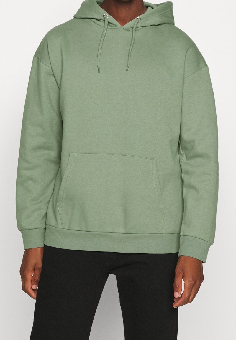 Homme portant un sweat à capuche vert clair uni avec poche avant et cordons, associé à un pantalon noir.