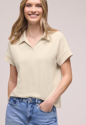 Mujer sonriente con cabello rubio lleva una camisa beige de manga corta con cuello y vaqueros azules con la mano derecha en un bolsillo.