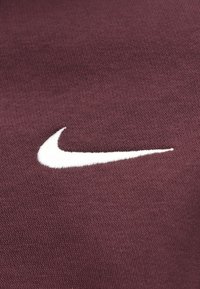 Material burgundiu cu o țesătură texturată prezintă un logo Nike swoosh alb brodat în partea dreaptă jos.