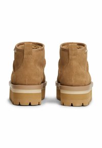 Botas tobilleras de ante marrón con una suela gruesa y estriada que presenta acentos en beige y crema. Incluye detalles de costura y una pestaña en la parte trasera.