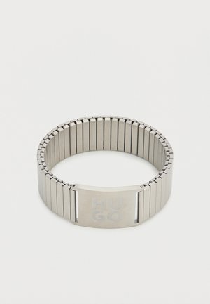Bracelet en métal avec un bracelet flexible, présentant une finition argent brossé. Gravé "HUGO" sur un fermoir rectangulaire. Design élégant et moderne.