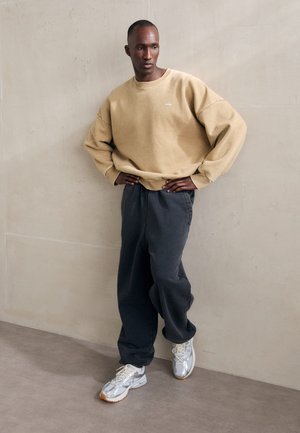 Homme portant un sweat-shirt beige oversize, un pantalon ample gris foncé et des baskets blanches, debout les mains sur les hanches contre un mur uni.
