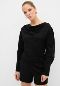 Schwarzes langärmeliges Oberteil mit einer drapierte Degolleté, aus glattem Stoff und mit feinen Falten an den Schultern. Kombiniert mit schwarzen Shorts.