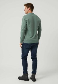 Hombre vistiendo un sudadera verde de manga larga, jeans azul oscuro y botas negras, de pie y de espaldas sobre un fondo claro liso.