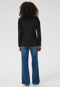 Göndör sötét hajú személy fekete zakót, kék bőrnadrágot és fekete magassarkú cipőt visel, háttal áll egy sima háttér előtt.